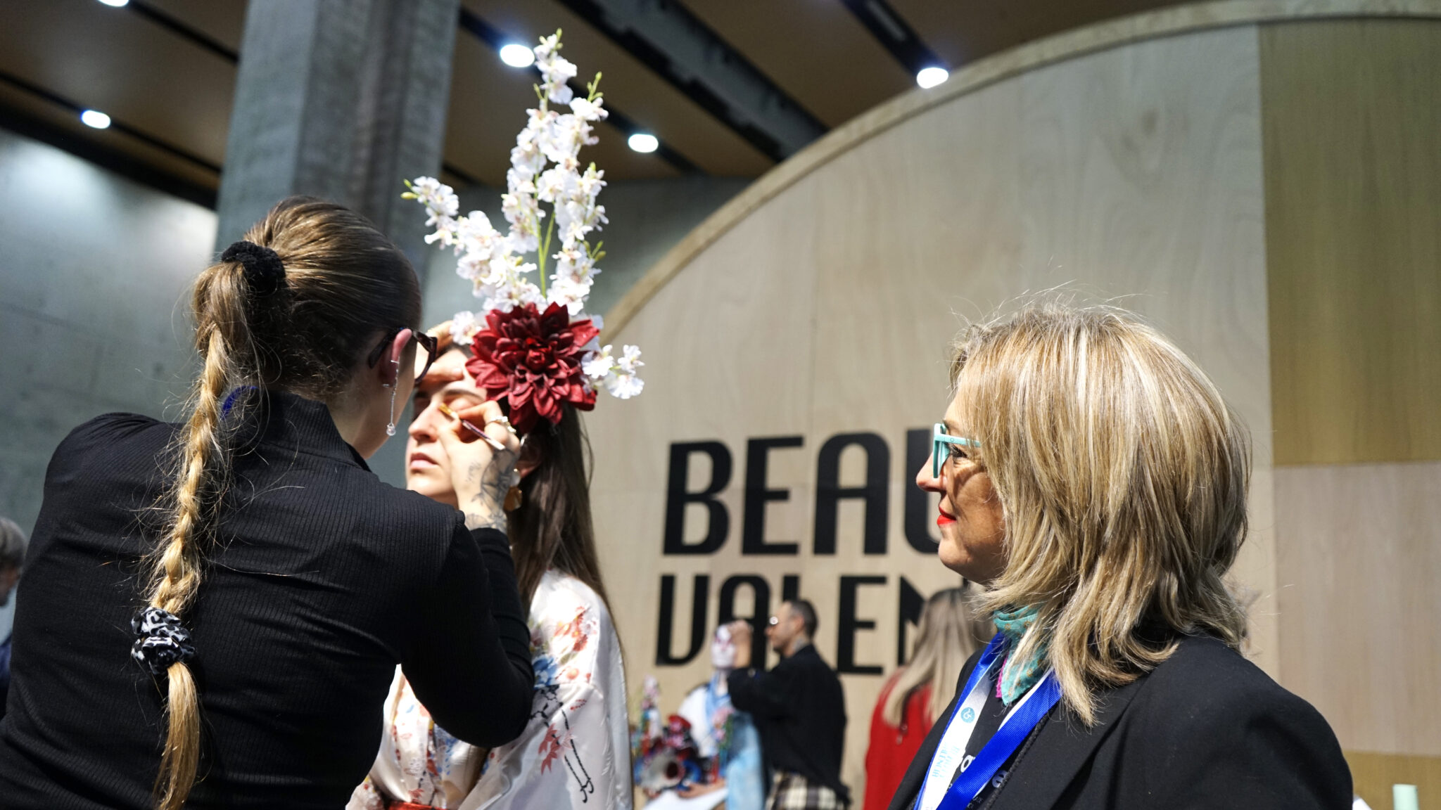 Una esteticista termina el maquillaje de una modelo con tocado floral mientras una socia de AVAPE observa, ante el rótulo de Beauty Valencia